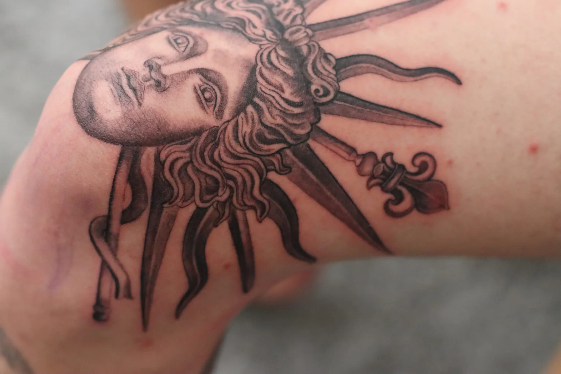 Black and grey tattoo of a classical face with curly hair and a sunburst halo on the arm near the elbow, Vienna