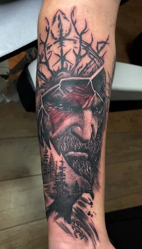 Black and grey forearm tattoo: a Viking with red war paint, an antler-and-rune crown, a raven and a pine forest