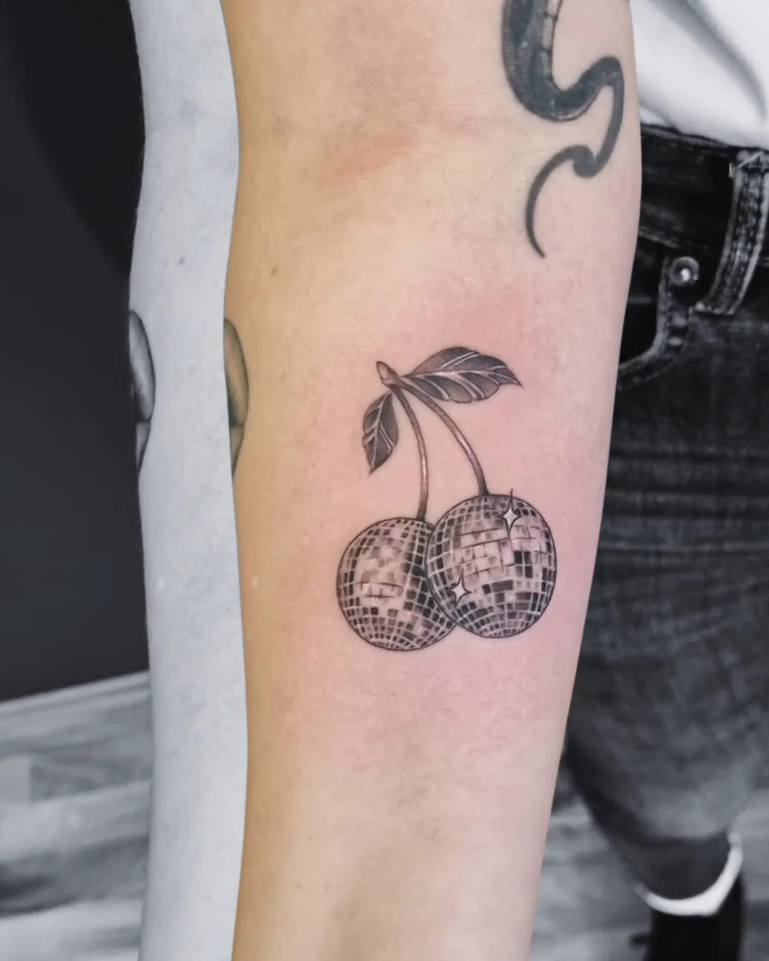 Fine line tattoo of two cherries as sparkling disco balls with leaves on the forearm, black and grey, tattoo studio Vienna