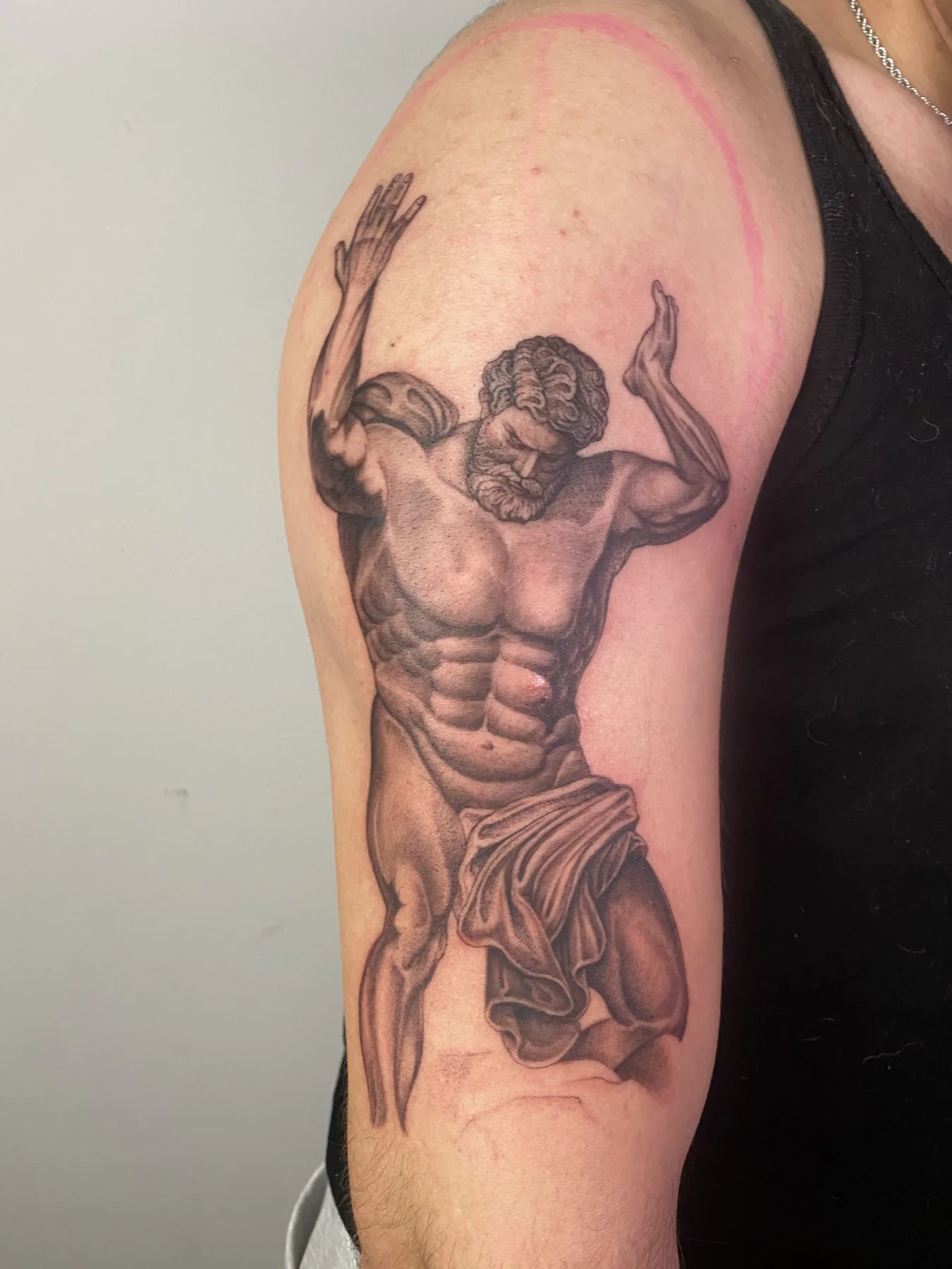 Realistic black and grey tattoo of an ancient Atlas statue with raised arms and drapery on the upper arm, tattoo Vienna