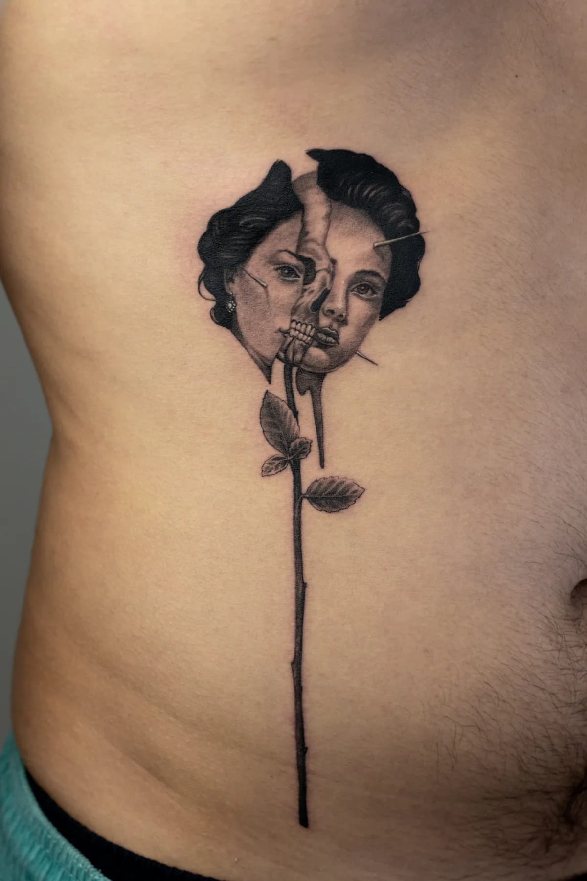 Fine black and grey tattoo on the ribs: a split woman's face revealing a skull, set on the long stem of a rose