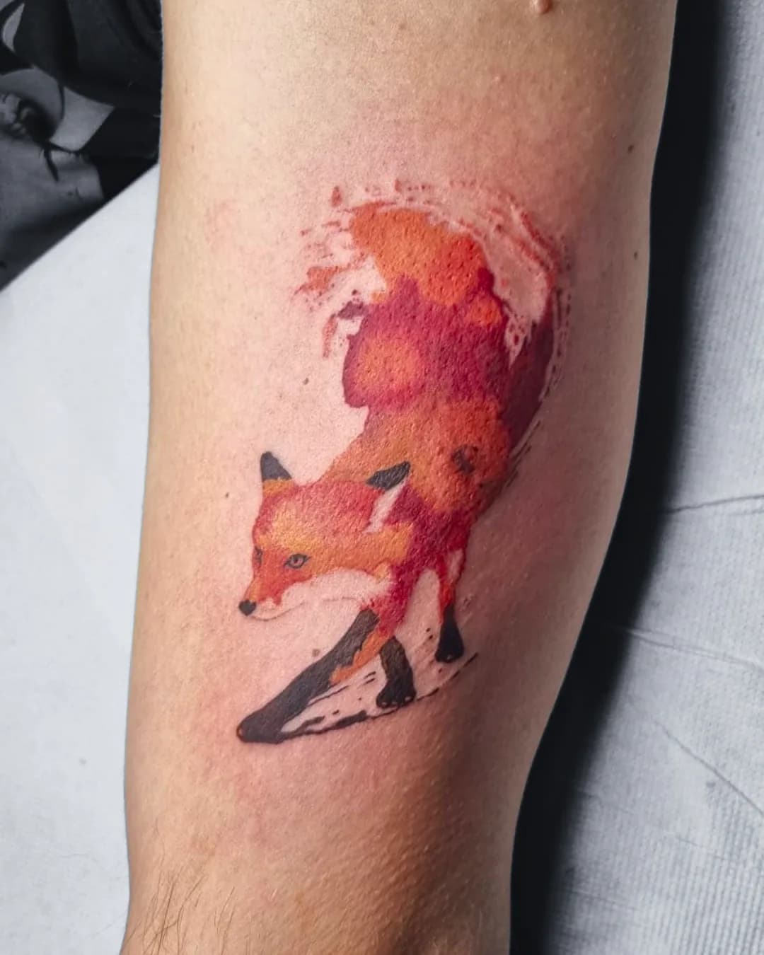 Watercolour tattoo of a fox in orange and red tones with black paws on the arm, aquarelle style, tattoo studio Vienna
