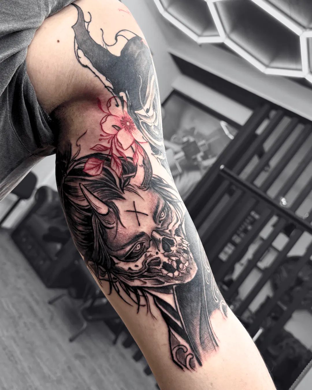 Black and grey arm tattoo: horned demon woman, half her face a skull, a cross on her forehead and a red flower, Vienna