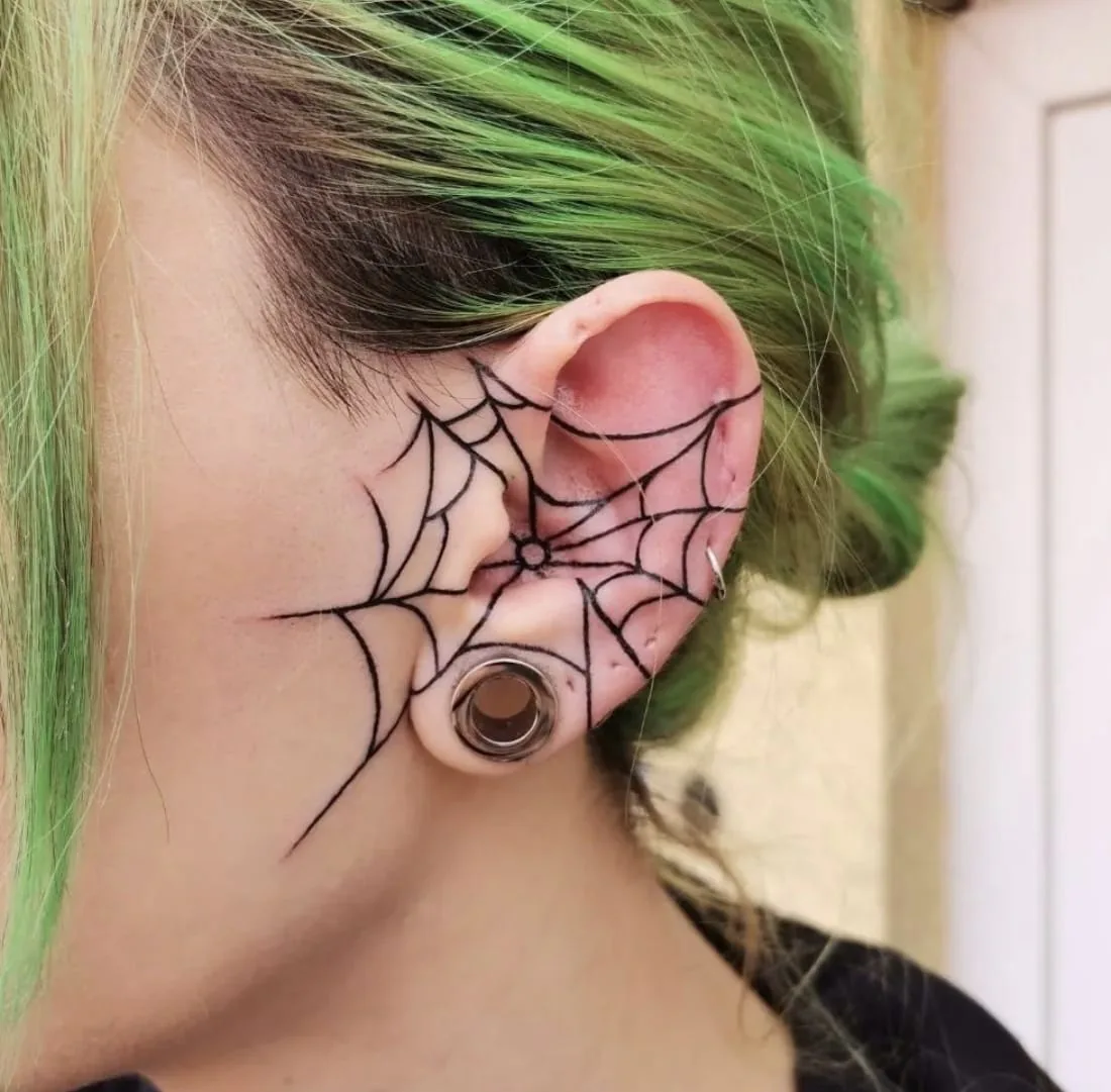 Fine line black spider web tattoo across the ear and cheek, next to an ear tunnel, tattoo studio Vienna