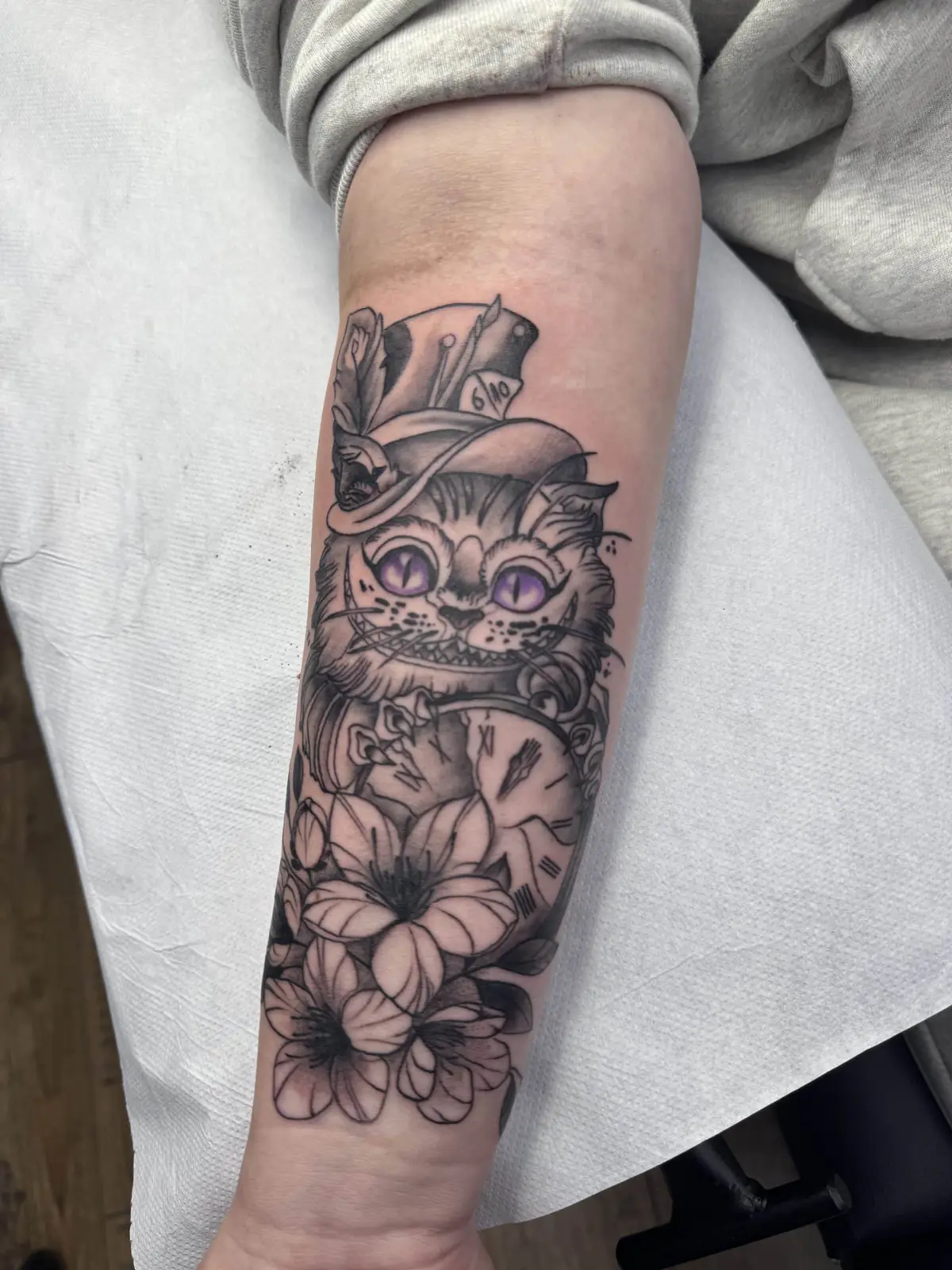 Tattoo of the Cheshire Cat with a top hat, violet eyes, a pocket watch and lilies on the forearm, tattoo Vienna