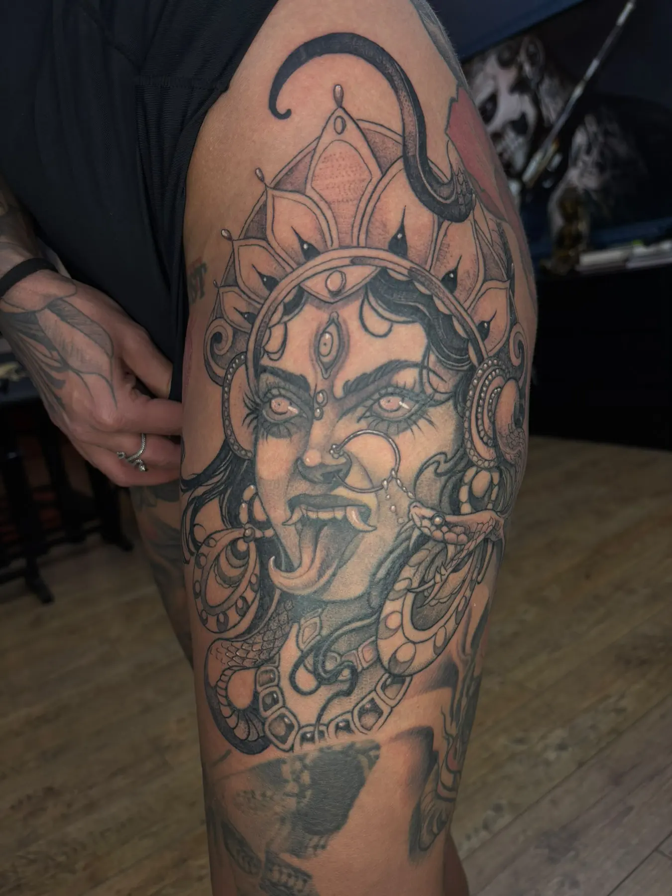 Illustrative black and grey tattoo of the goddess Kali with a third eye, outstretched tongue, snakes and a crown on the thigh