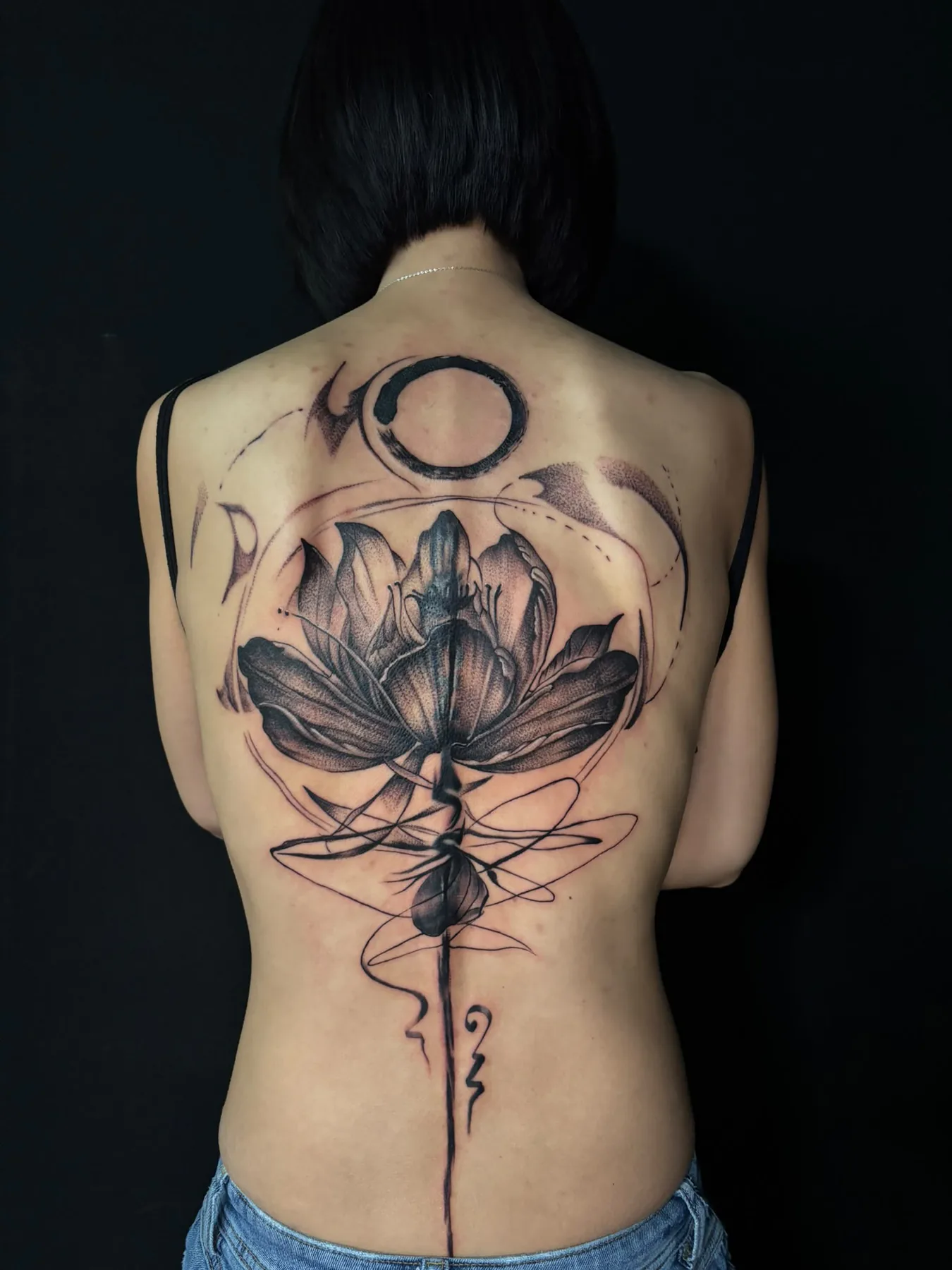 Large black and grey back tattoo: a lotus flower with an ensō circle, abstract lines and one line running down the spine