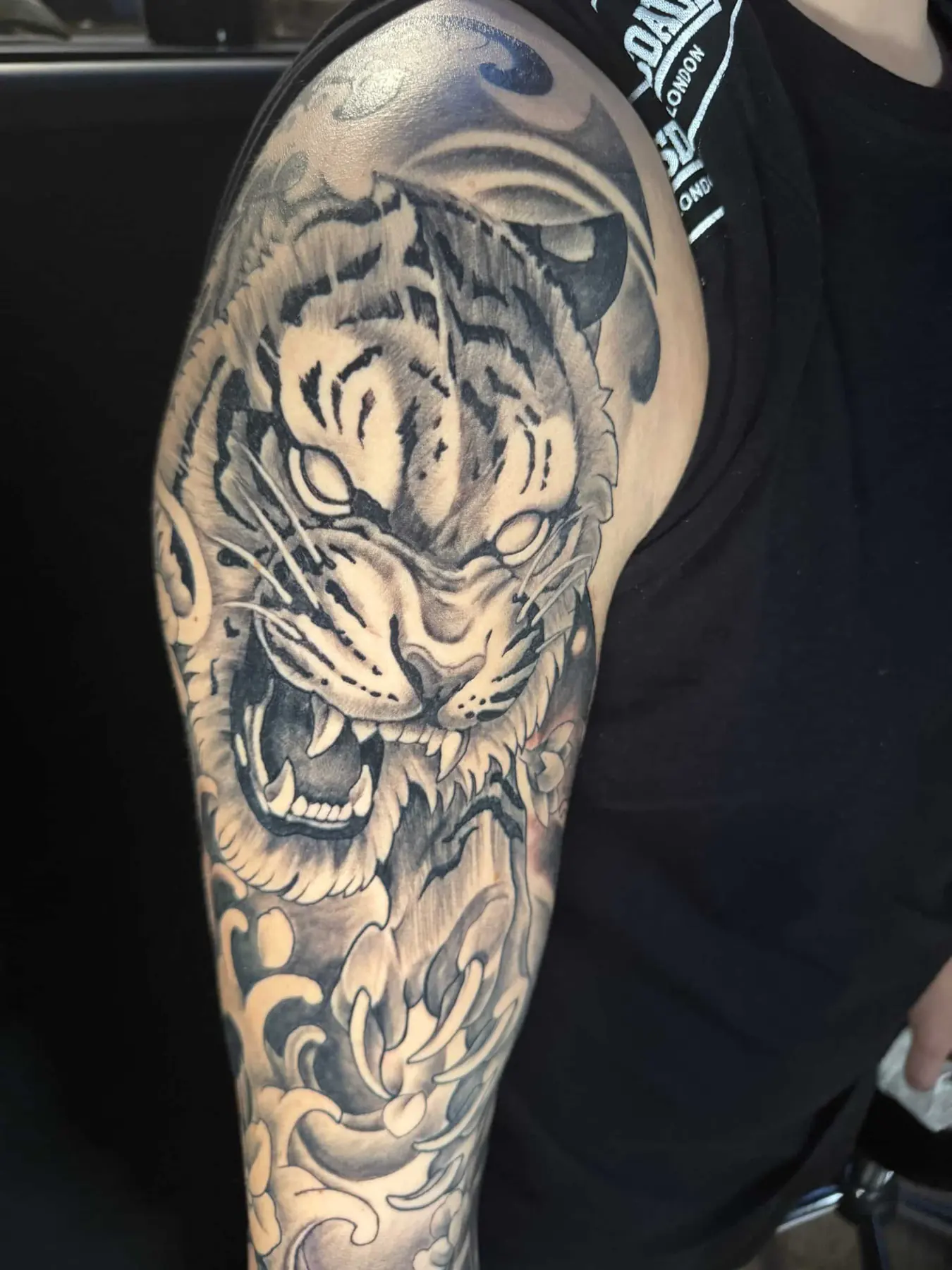 Japanese-style black and grey tattoo: a roaring tiger with chrysanthemums and waves on the upper arm and shoulder, Vienna