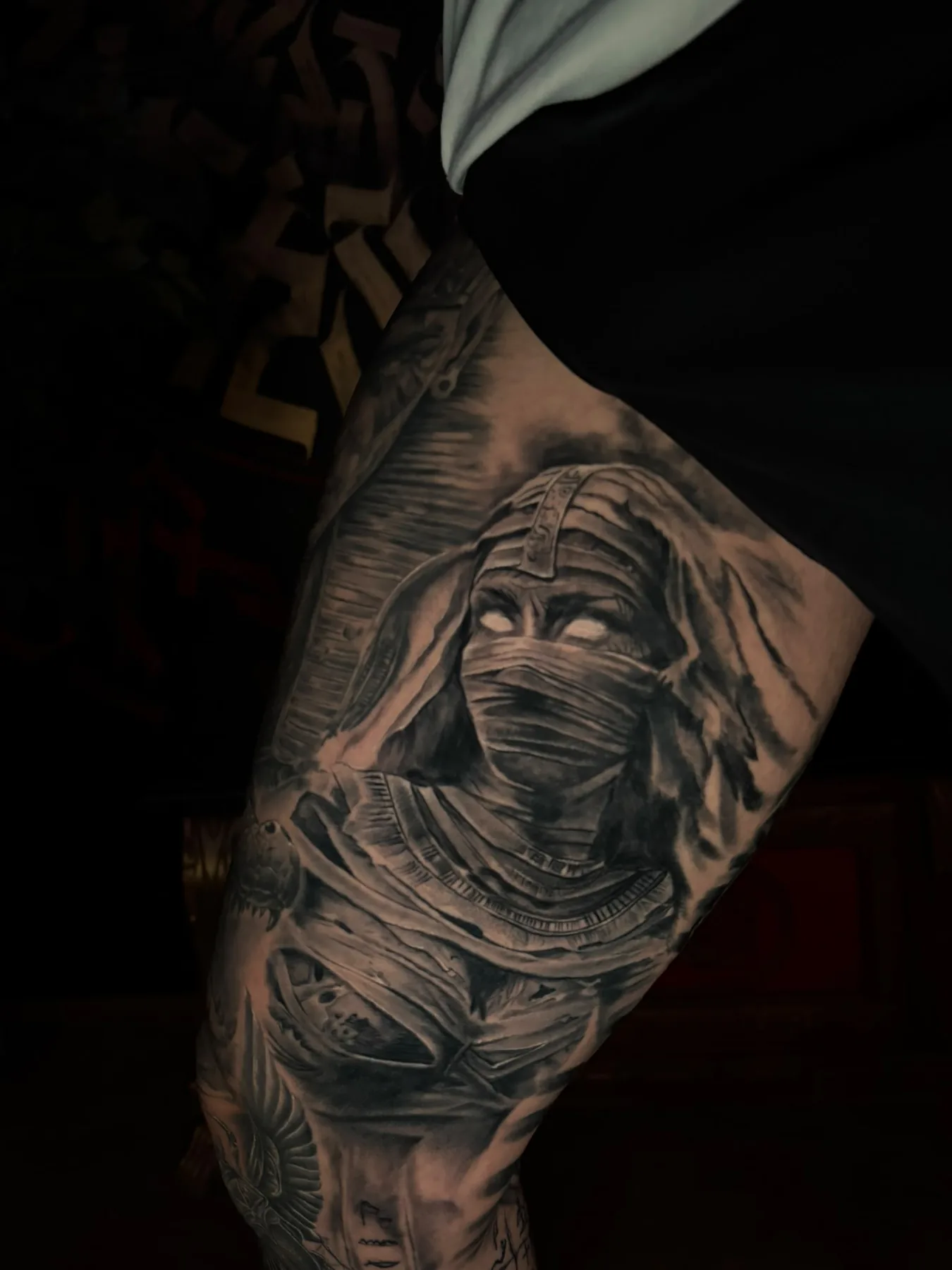 Black and grey realism tattoo of a veiled mummy with glowing white eyes on the thigh, part of an Egyptian leg sleeve, Vienna