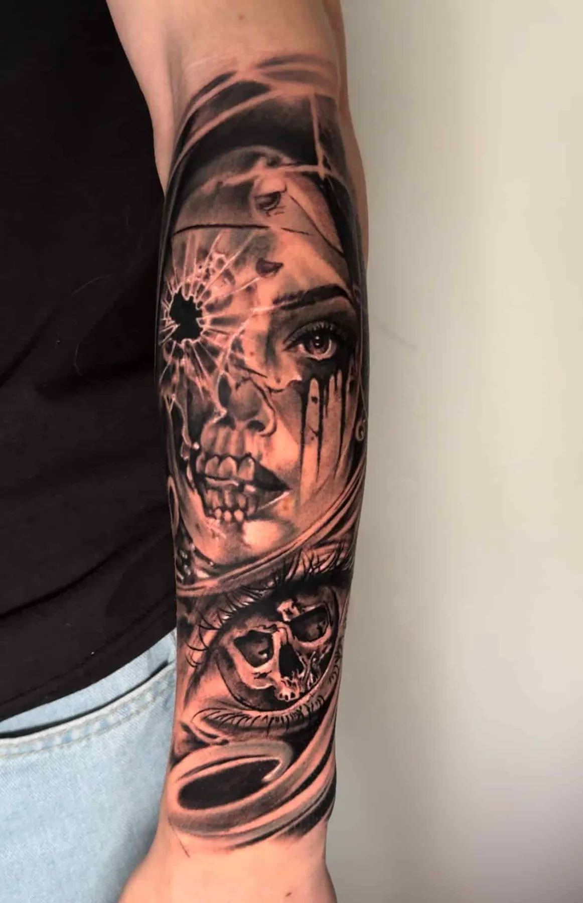 Realistic black and grey forearm tattoo: a woman's face shattering around a bullet hole into a skull, with an eye below