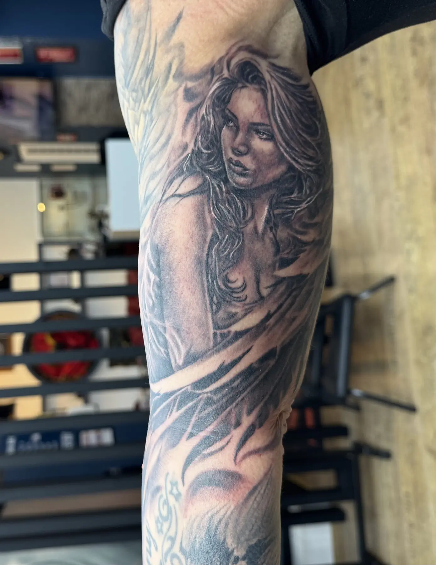 Realistic black and grey portrait of a woman with long wavy hair and wings on the upper arm, tattoo studio Vienna