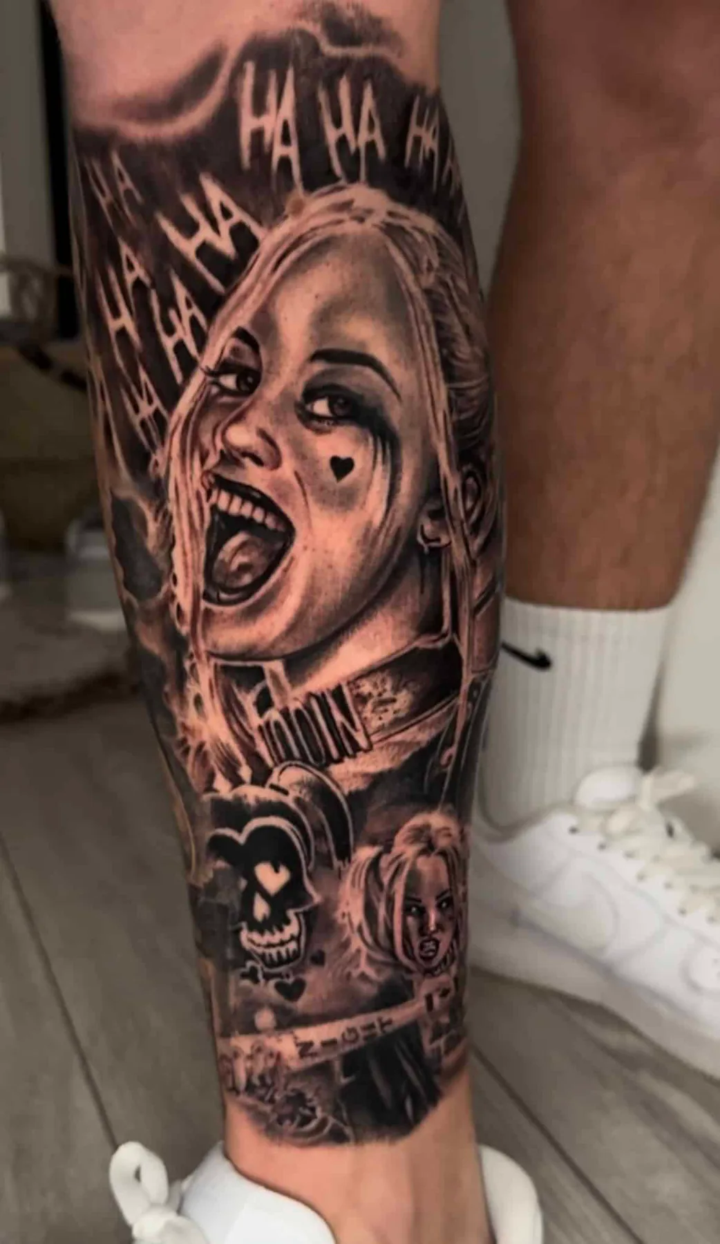 Realistic black and grey portrait of Harley Quinn laughing with "HA HA" lettering and a jester skull on the calf, Vienna