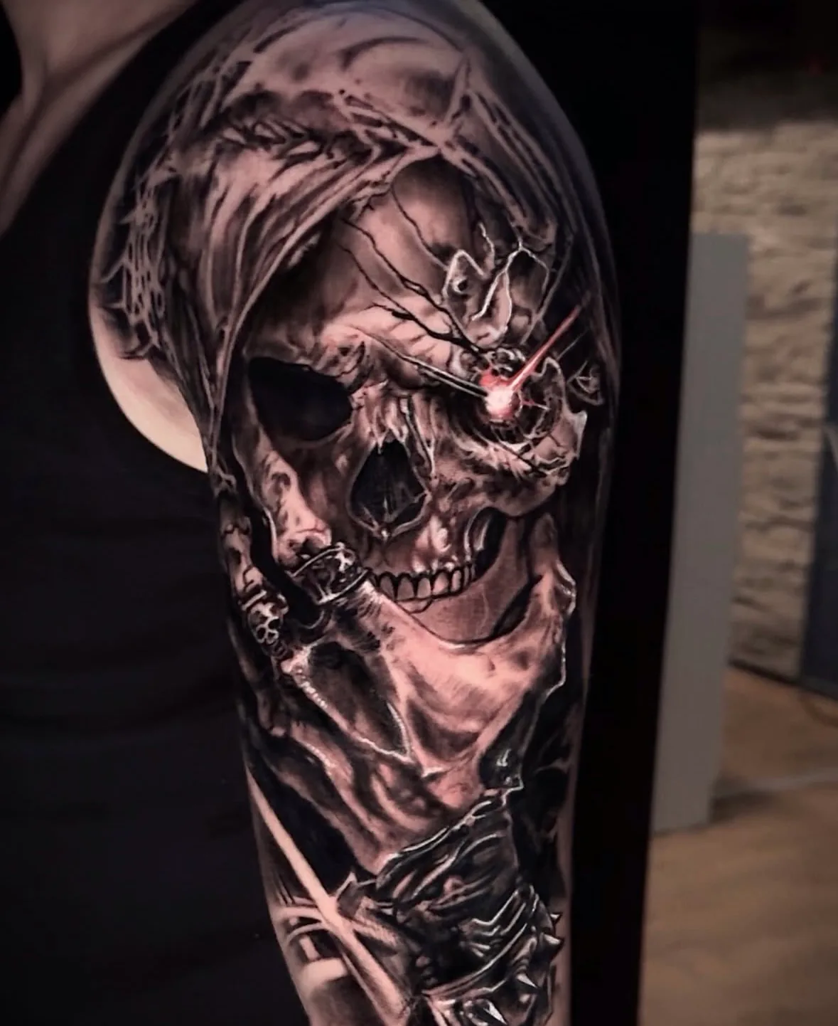 Black and grey horror tattoo: a hooded grim reaper skull with a red light beam in its eye on the upper arm, tattoo Vienna