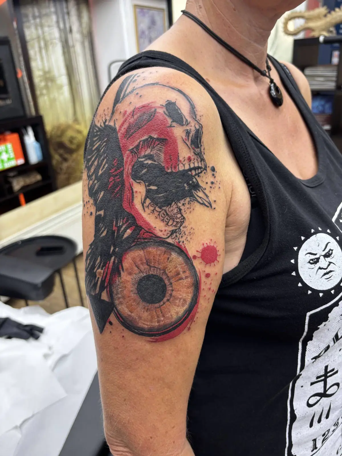 Trash polka tattoo on the shoulder: a raven and a skull in black with red splashes and a realistic brown eye
